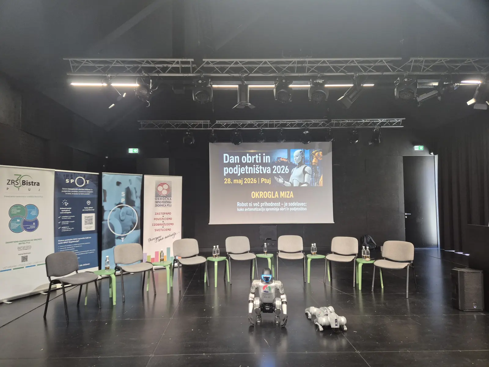 Prizorišče okrogle mize: stoli v polkrogu, projekcija »Dan obrti in podjetništva 2026« ter humanoidni in štirinožni robot.