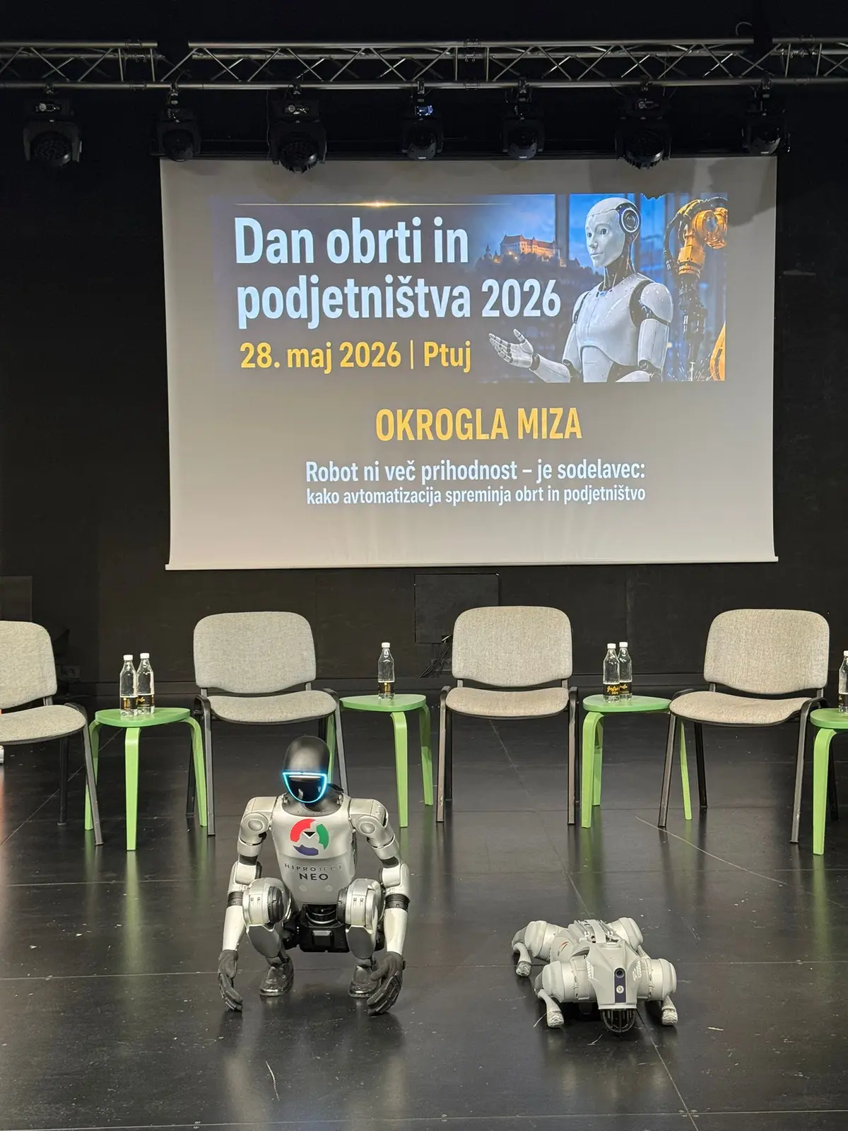 Humanoidni robot HiProject NEO in štirinožni robot pred prosojnico okrogle mize.