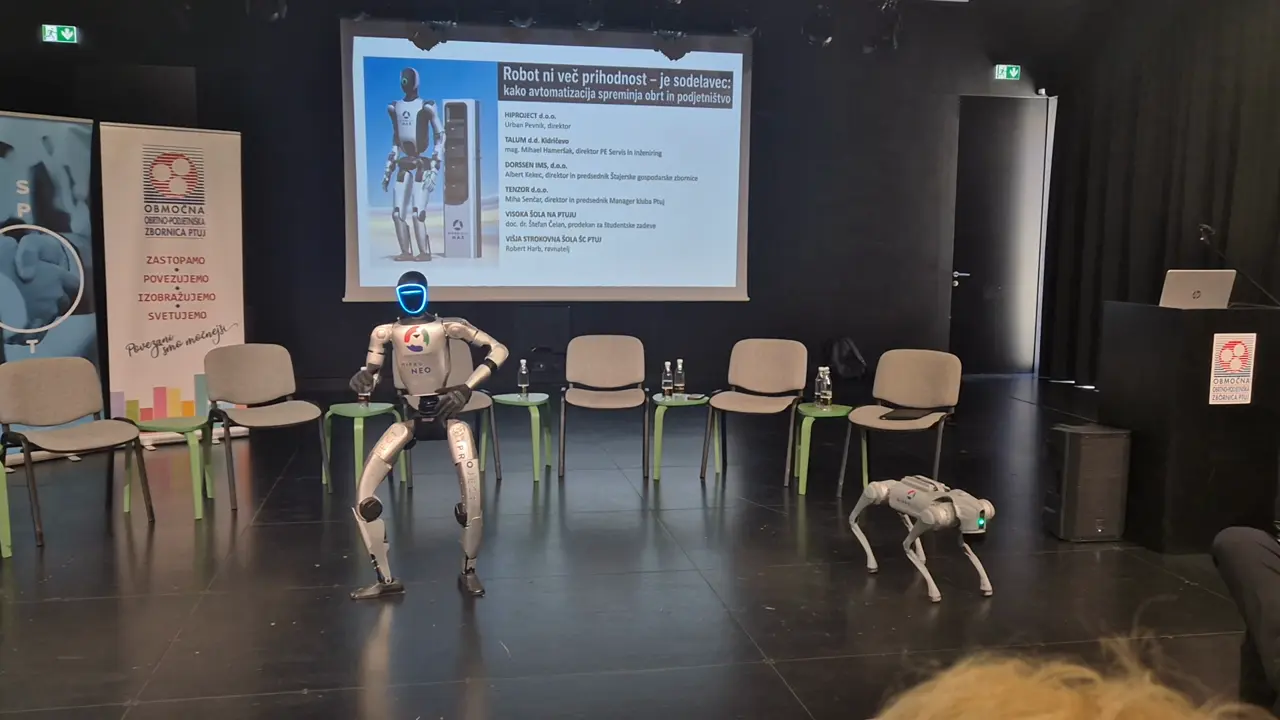 Humanoidni in štirinožni robot na odru pred prosojnico okrogle mize.